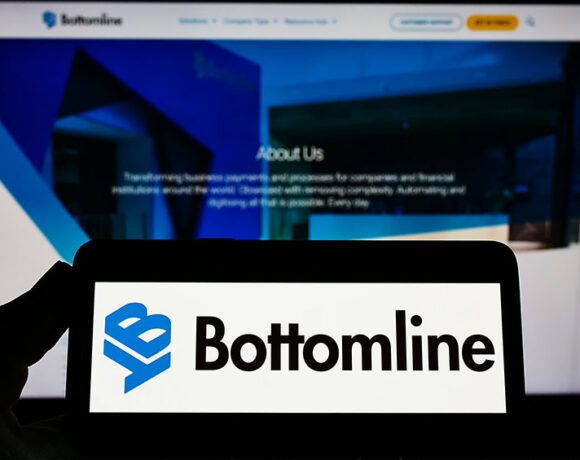 A person holding a smartphone that displays the logo of Bottomline in front of the official website of Bottomline on account of Nexus Systems acquisition.