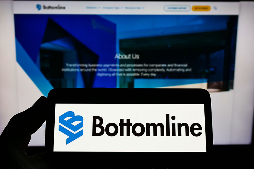 A person holding a smartphone that displays the logo of Bottomline in front of the official website of Bottomline on account of Nexus Systems acquisition.