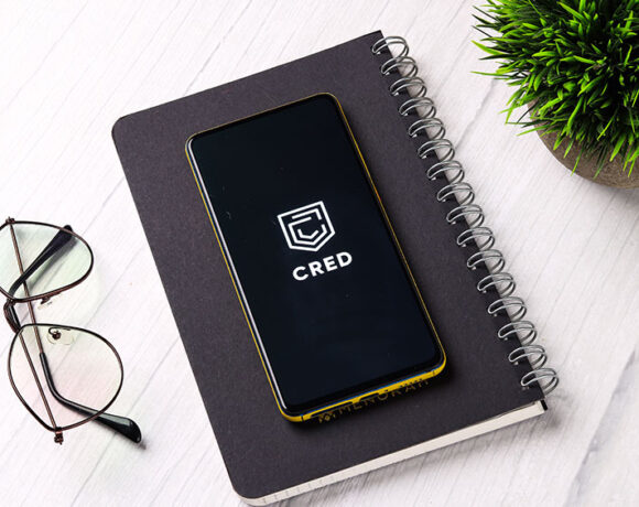 A smartphone with a logo Cred, is kept on a notebook near a eyeglasses.
