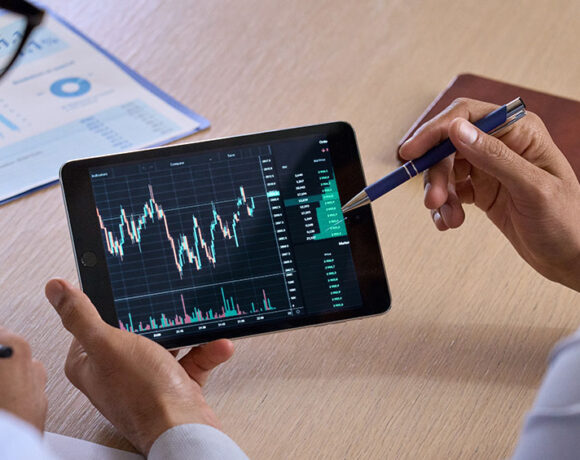 Two business professionals consulting and analyzing a crypto business chart on a smart phone with a pen in their hand, pie chart paper can be seen in the vicinity.