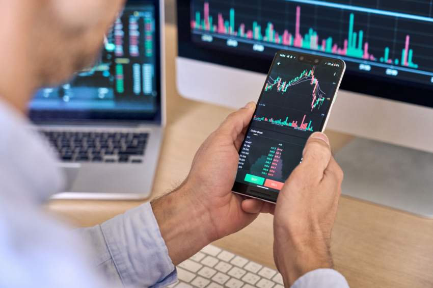 An over-shoulder view of a crypto investor using a smartphone app to analyze financial data, checking the stock market price, and checking the online trading platform.